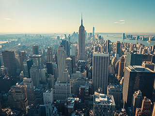 Aerial view of a sprawling cityscape with densely packed buildings, streets, and a backdrop of hills and the sea in the distance.